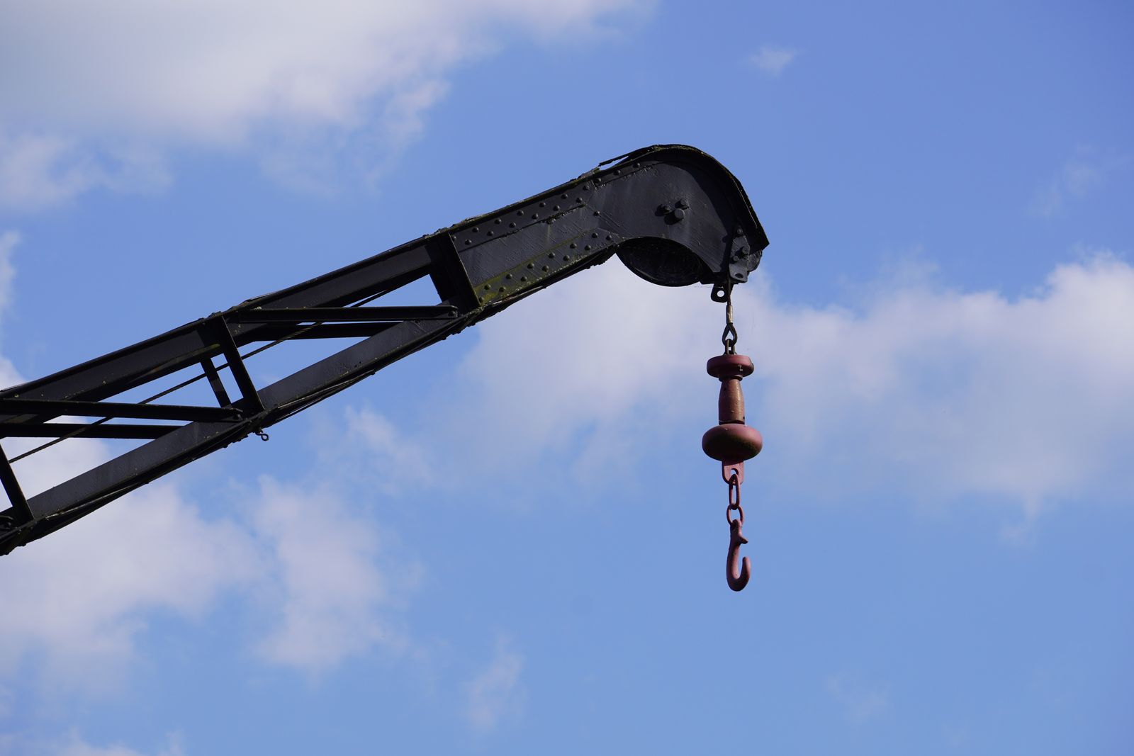 The black boom tip of a knuckle-boom crane with its hook block hanging against a blue sky with white clouds