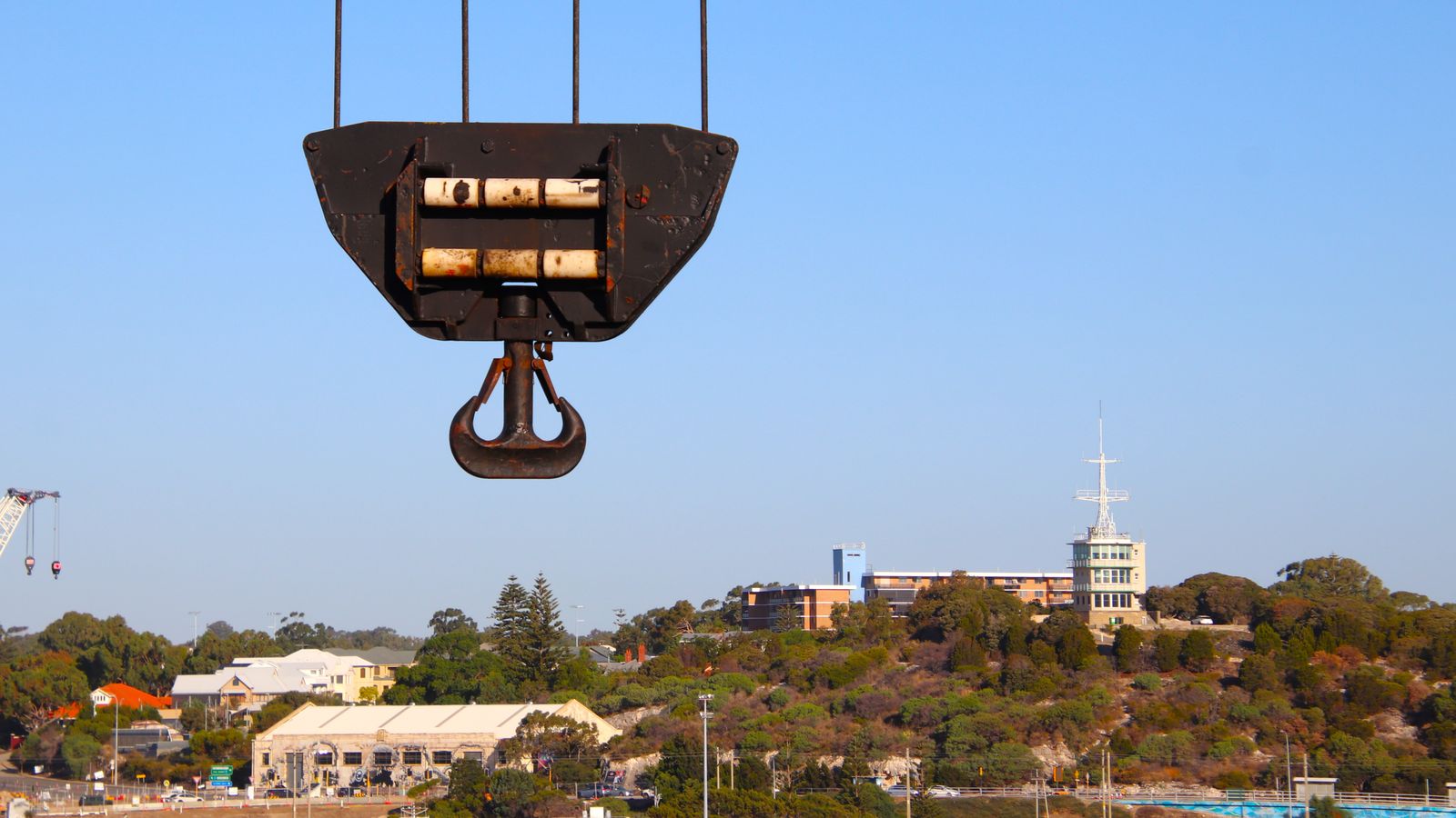 A weathered crane hook block hanging on wire ropes against a clear blue sky above a hillside town