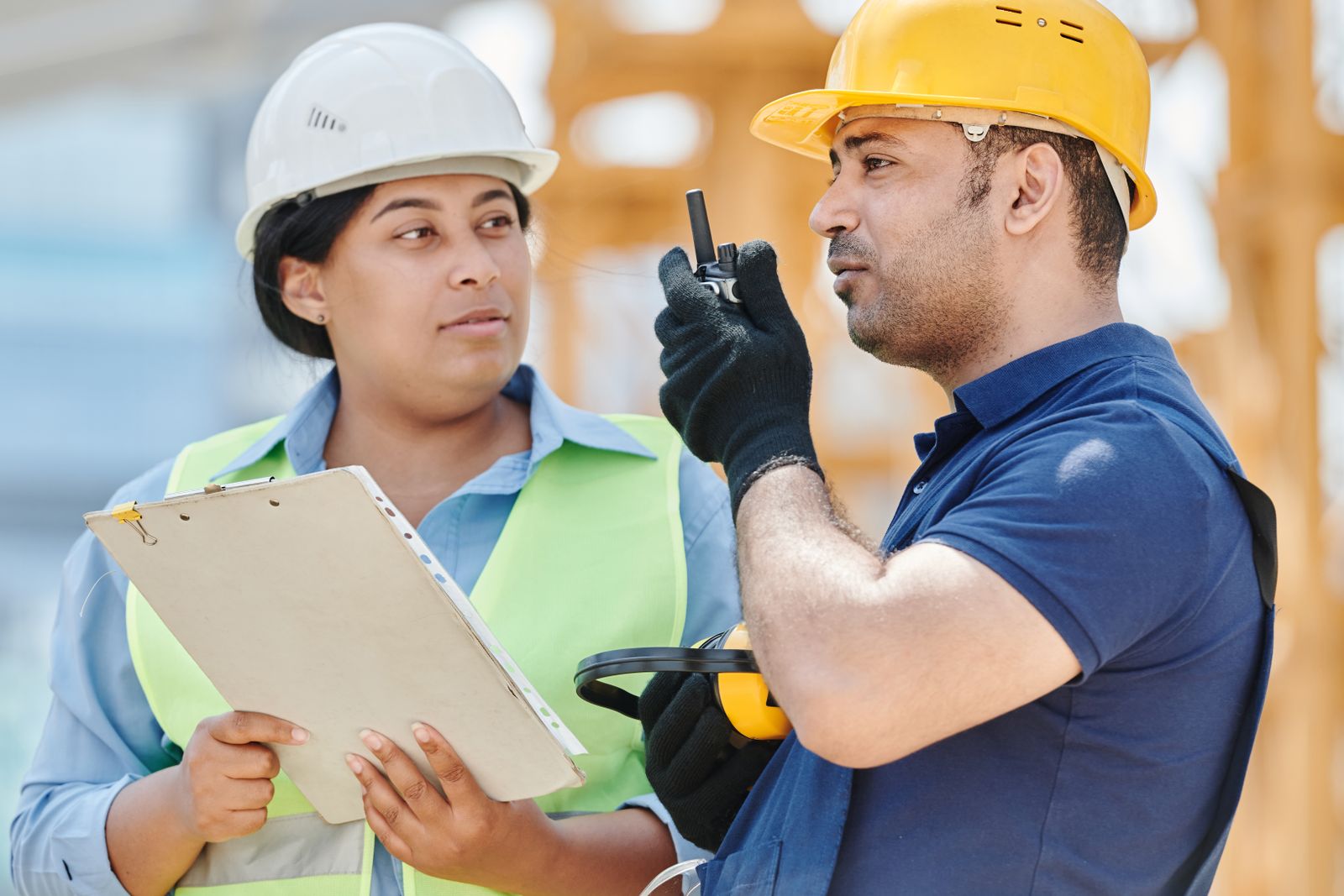 A worker in a white hard hat holding a clipboard next to a man in a yellow hard hat talking on a handheld radio