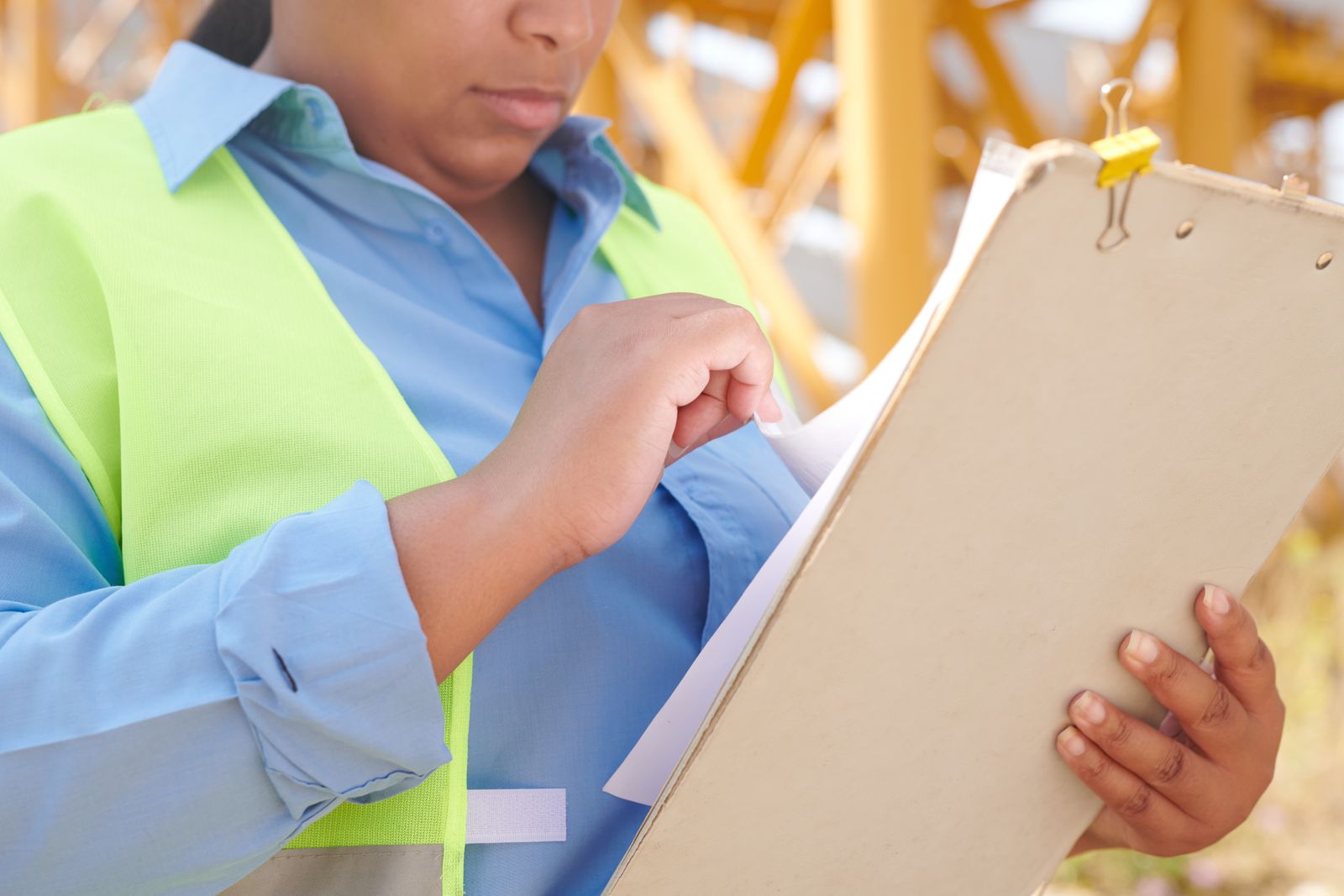 A worker in a hi-vis vest and blue shirt flipping through papers on a clipboard in front of a yellow steel frame