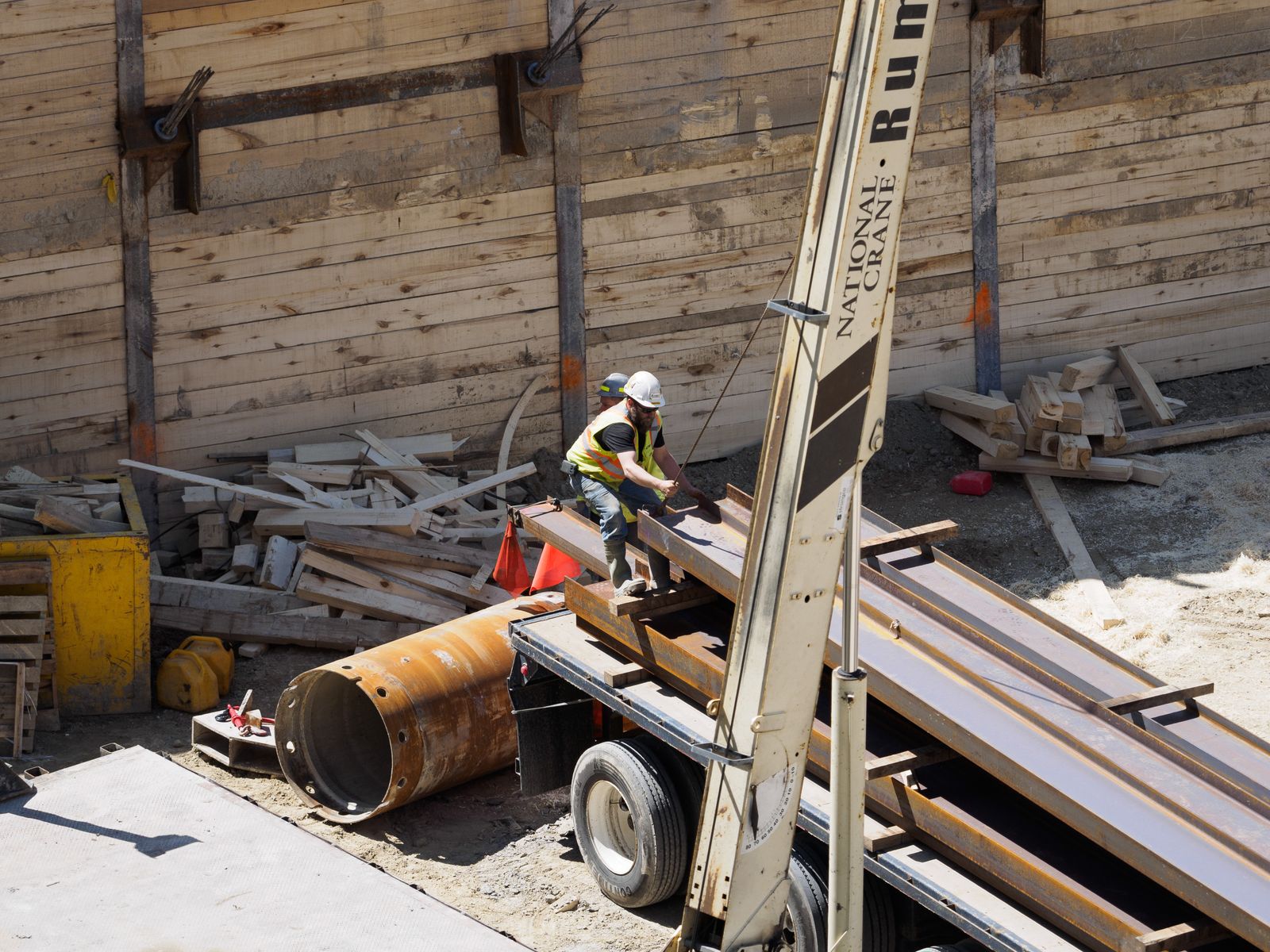 A worker in a hard hat and hi-vis vest rigging steel beams on a flatbed truck beside a boom truck crane at an excavation
