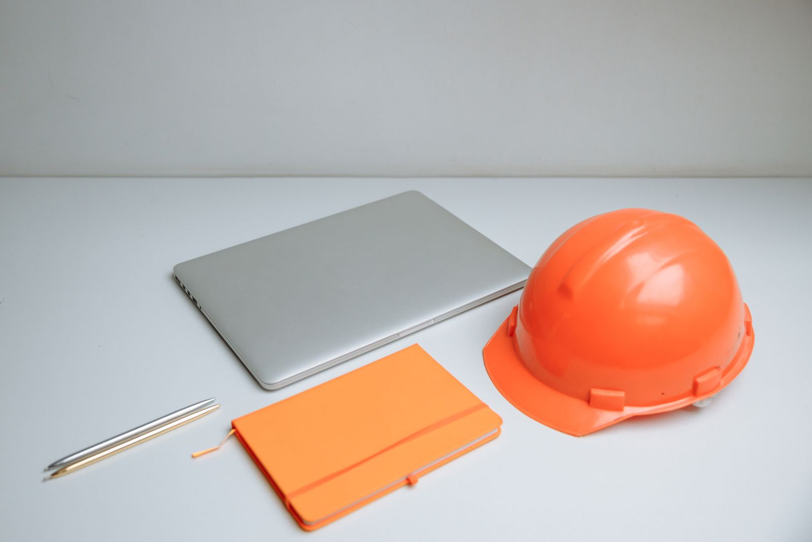 A closed laptop, an orange notebook, two pens and an orange hard hat on a plain white desk