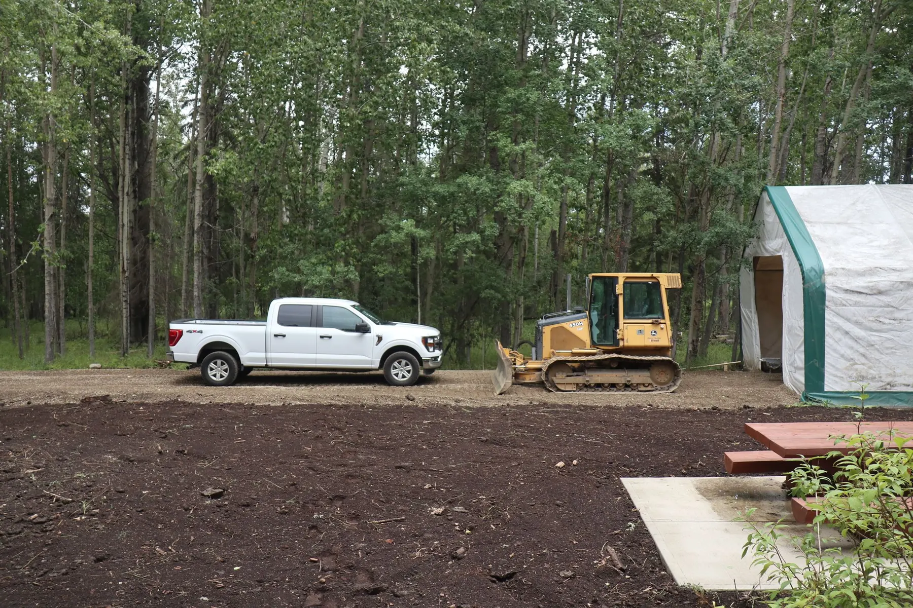 White pickup truck and bulldozer parked by a forest with a shelter nearby.